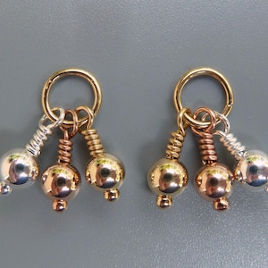 May include: A pair of earrings with a gold hoop and three dangling charms. The charms include a silver, copper, and gold-colored sphere. The earrings are displayed against a gray background.