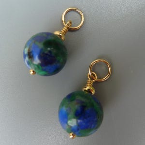 May include: Two round earrings with a gold-colored loop and accents. The earrings feature a blue and green marbled stone. The earrings are displayed against a gray background.