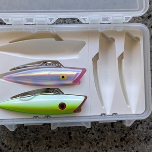 May include: A clear plastic tackle box with a white interior holds fishing lures. Two lures are visible: one iridescent and pink, the other green and red. Also included are white plastic blades.