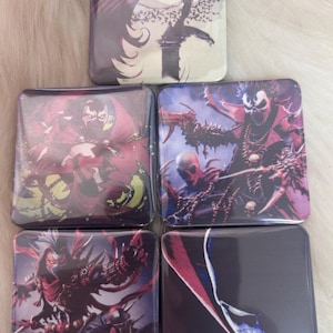 May include: Five square-shaped buttons with different images of the comic book character Spawn. The buttons feature a variety of poses and backgrounds, including a cityscape, a moonlit sky, and a fiery inferno.