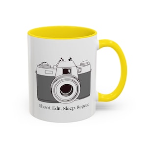 Könnte beinhalten: Eine weiße Keramiktasse mit gelbem Rand und Henkel. Die Tasse zeigt eine Schwarzweißillustration einer Vintage-Kamera und den Text "Shoot. Edit. Sleep. Repeat."