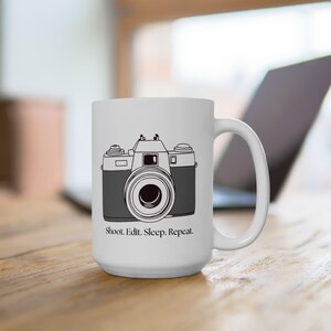 Könnte beinhalten: Eine weiße Keramiktasse mit einer Schwarzweißillustration einer Kamera und dem Text "Shoot. Edit. Sleep. Repeat."