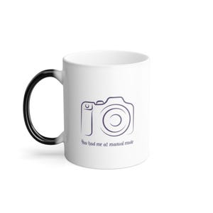 May include: White ceramic coffee mug with a black handle. The mug has a black outline of a camera with the text "You had me at manual mode" printed on the front.