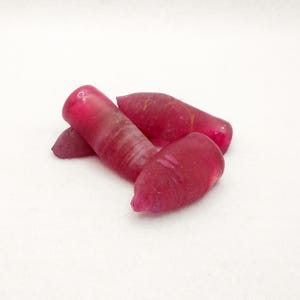 Puede incluir: Cuatro piedras preciosas de rubí en bruto sin cortar. Las piedras preciosas son de un color rojo intenso y saturado con una apariencia ligeramente translúcida. Las formas son irregulares, con extremos redondeados y una textura natural sin pulir.