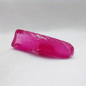 May include: A raw, uncut ruby gemstone. The ruby is a vibrant, translucent pink color with a slightly rough, textured surface. The gemstone is elongated and has a natural, organic shape, resting on a white surface.