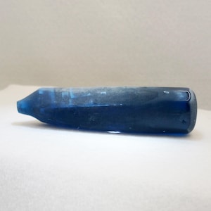 Puede incluir: Un tapón de botella azul oscuro con un extremo redondeado y una punta cónica. El tapón tiene un aspecto esmerilado y está hecho de un material sólido. El objeto está acostado horizontalmente sobre una superficie blanca.