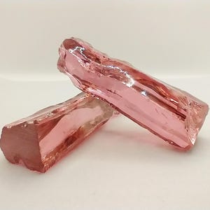 Puede incluir: Dos piedras de cristal rosa facetadas. Las piedras son translúcidas y tienen un ligero tono rojizo.