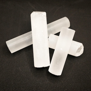 Può includere: Quattro bacchette di selenite bianche, anche conosciute come bastoncini di selenite, sono disposte a forma di croce su una superficie nera. Le bacchette sono lisce e traslucide, con un aspetto leggermente nuvoloso.