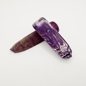 以下が含まれることがあります： 2つの紫色の宝石。1つは粗い自然な仕上げ、もう1つは滑らかで磨かれた仕上げ。磨かれた宝石は銀色の光沢があります。