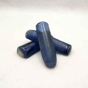 May include: Three dark blue glass rods are arranged on a white surface. The rods have a cylindrical shape with rounded ends. The glass appears to have a slight gradient in color, transitioning from a darker blue to a lighter shade.