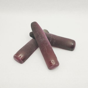 May include: Three tumbled, elongated gemstones in varying shades of deep red-purple are arranged on a white surface. The stones have rounded ends and a smooth, polished appearance, with subtle variations in color and texture.