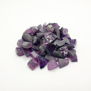 Puede incluir: Un montón de virutas de piedras preciosas púrpuras, probablemente amatista o zafiro, con una variedad de formas y tamaños. Las virutas están dispuestas en un montón suelto sobre un fondo blanco.