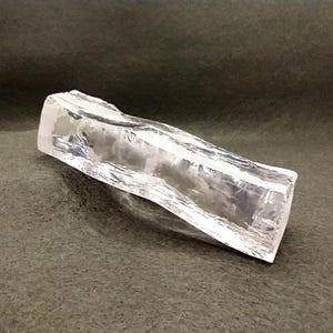 May include: A clear, rectangular crystal with a rough, textured surface. The crystal is transparent, allowing light to pass through, and is set against a dark background. The crystal's shape is elongated, with a slightly irregular form.