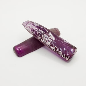 Op de afbeelding: Twee ruwe paarse edelstenen, een is langer en heeft een puntiger uiteinde, de andere is korter en heeft een ronder uiteinde.