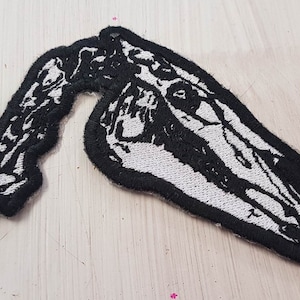 May include: Black and white embroidered patch of a skull with a skeletal neck. The skull is facing to the right and has a detailed design.
