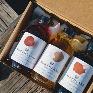 May include: A gift box containing three glass bottles of Northland Vinegars. The bottles have black caps and white labels with the brand name and flavor. The box is filled with white paper and sits on a wooden surface.