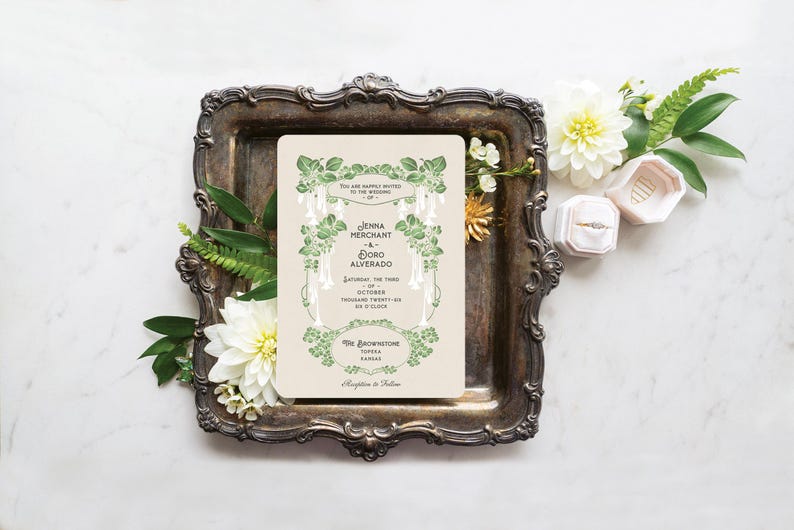 K&ouml;nnte beinhalten: Eine Hochzeitseinladung mit gr&uuml;nen floralen Akzenten und Text, platziert auf einem Vintage-Silbertablett. Die Einladung ist von wei&szlig;en Blumen, Gr&uuml;n und zwei Ringboxen umgeben. Die Einladung enth&auml;lt die Namen Jenna Merchant und Doro Alvarado.