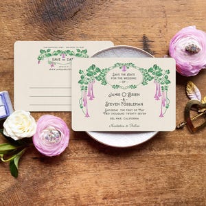 K&ouml;nnte beinhalten: Hochzeitseinladungs-Set mit einer cremefarbenen Karte mit gr&uuml;nem und rosa floralem Design, einer Ringbox, Blumen und einem dekorativen Herz auf einer Holzoberfl&auml;che. Die Karte tr&auml;gt die Aufschrift "Save the Date".