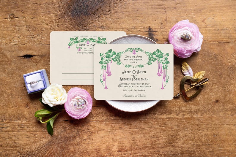 K&ouml;nnte beinhalten: Hochzeitseinladungs-Set mit einer cremefarbenen Karte mit gr&uuml;nem und rosa floralem Design, einer Ringbox, Blumen und einem dekorativen Herz auf einer Holzoberfl&auml;che. Die Karte tr&auml;gt die Aufschrift "Save the Date".