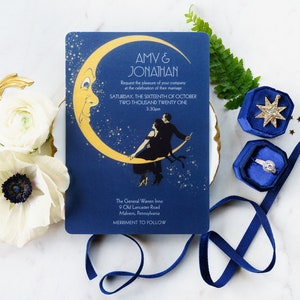 May include: A navy blue wedding invitation with gold foil accents features a couple dancing on a crescent moon with stars in the background. The invitation text reads "AMV & JONATHAN" and includes the date and time of the wedding. The invitation is on a white surface with blue ribbon, a blue ring box, and gold stars.