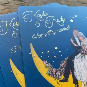 Puede incluir: Una invitaci&oacute;n de boda azul con letras doradas. La invitaci&oacute;n presenta a una pareja sentada en una luna creciente con estrellas en el fondo. El texto dice "Kayla & Rudy Are getting married!"