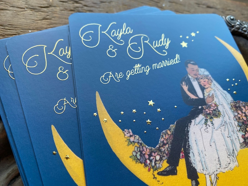 Puede incluir: Una invitaci&oacute;n de boda azul con letras doradas. La invitaci&oacute;n presenta a una pareja sentada en una luna creciente con estrellas en el fondo. El texto dice "Kayla & Rudy Are getting married!"