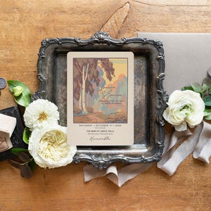 May include: A vintage-style wedding invitation with a landscape illustration, displayed on a decorative silver tray. The invitation is accompanied by white flowers, a gray envelope, and wax seals, all arranged on a wooden surface.