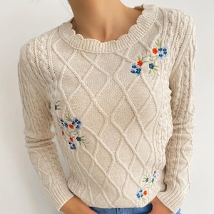Könnte beinhalten: Cremefarbener Zopfstrickpullover mit Rautenmuster und Blumenstickerei in Blau und Rot. Der Pullover hat einen wellenförmigen Ausschnitt und lange Ärmel. Die Person trägt eine blaue Jeans.