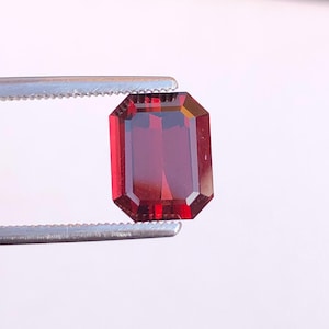 May include: A faceted, rectangular-cut, deep red gemstone is held by a pair of silver tweezers. The gem has a polished surface and a rich, saturated color. The gemstone is set against a plain white background.