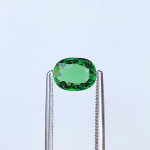 Könnte beinhalten: Ein lebendiger grüner Edelstein mit ovalem Schliff, gehalten von einer silbernen Pinzette. Der Edelstein zeigt eine satte, smaragdgrüne Farbe und eine polierte Oberfläche, die das Licht reflektiert. Der Hintergrund ist ein sauberes, neutrales Weiß.