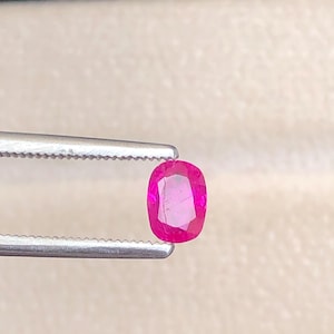 Könnte beinhalten: Ein lebendiger, oval geschliffener Rubin, gehalten von einer silbernen Pinzette. Der Rubin zeigt eine satte, gesättigte rosa Farbe mit sichtbaren inneren Facetten. Die Pinzette ist scharf fokussiert und hebt die Klarheit und Brillanz des Edelsteins hervor.