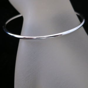 Peut inclure: Un fin bracelet jonc argenté brillant. Le bracelet circulaire semble être en métal. Le fond est noir et le bracelet repose sur une surface de couleur claire.