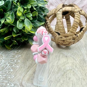 May include: A pink and white beaded bracelet with a pink ribbon charm. The bracelet features various shapes of beads, including round, cube, and heart-shaped, with silver accents. The ribbon charm is a symbol of breast cancer awareness.