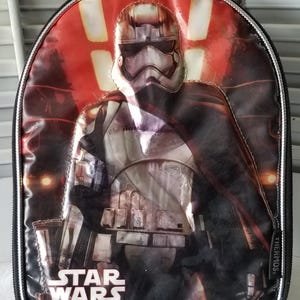 May include: Black lunch bag featuring a Star Wars design. The bag has a graphic of a Stormtrooper with a red and black background. The words "STAR WARS" are printed at the bottom of the bag.
