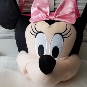Puede incluir: Bolso tote de felpa con forma de Minnie Mouse. El bolso presenta un lazo rosa con lunares blancos, orejas negras y un asa rosa. La cara es de color rosa claro con ojos negros y una nariz negra.