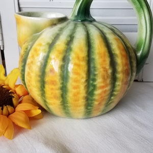 May include: A ceramic pitcher with a pumpkin design. The pitcher is primarily yellow and orange with green vertical stripes. It has a green handle and a small spout. A yellow sunflower is in the foreground.