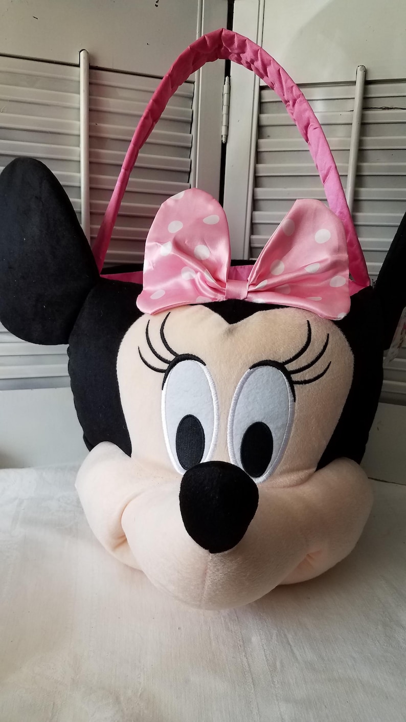 Puede incluir: Bolso tote de felpa con forma de Minnie Mouse. El bolso presenta un lazo rosa con lunares blancos, orejas negras y un asa rosa. La cara es de color rosa claro con ojos negros y una nariz negra.