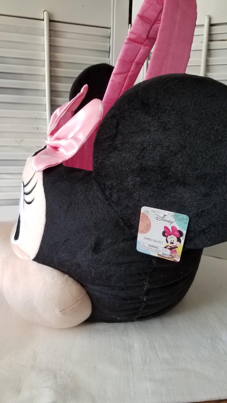 Puede incluir: Una cesta jumbo de Minnie Mouse de peluche en negro, rosa y blanco. La cesta tiene forma de cabeza de Minnie, con grandes orejas negras, un lazo rosa y orejas rosas. Una etiqueta de Disney est&aacute; adjunta.