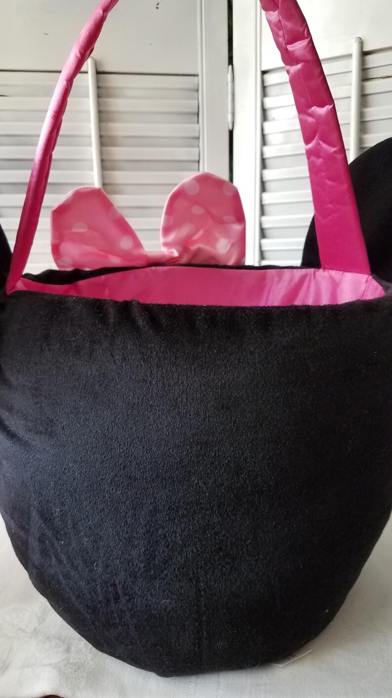 Puede incluir: Bolso tote de felpa negra con forro y asas de sat&eacute;n rosa. El bolso presenta un lazo rosa con lunares blancos. Las asas son de color rosa brillante. El bolso est&aacute; dise&ntilde;ado para parecerse a la cabeza de un personaje.