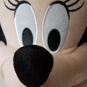 Puede incluir: Primer plano de la cara de peluche de Minnie Mouse. El personaje tiene una nariz negra, ojos blancos grandes con pupilas negras y largas pesta&ntilde;as negras. Un lazo rosa con lunares blancos est&aacute; en la parte superior de la cabeza. El peluche est&aacute; hecho de tela suave y clara.
