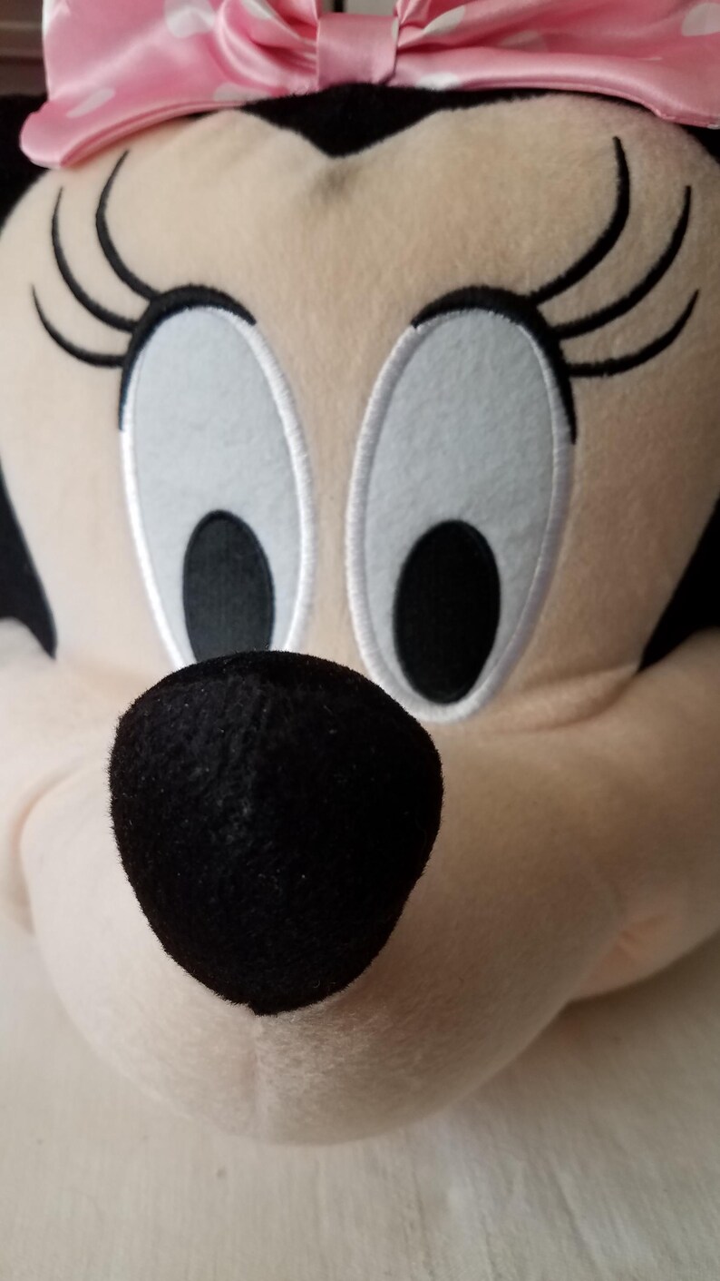 Puede incluir: Primer plano de la cara de peluche de Minnie Mouse. El personaje tiene una nariz negra, ojos blancos grandes con pupilas negras y largas pesta&ntilde;as negras. Un lazo rosa con lunares blancos est&aacute; en la parte superior de la cabeza. El peluche est&aacute; hecho de tela suave y clara.