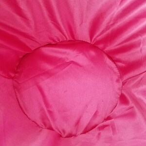 Puede incluir: Primer plano de un elemento de tela rosa brillante, circular, posiblemente un coj&iacute;n o parte de un textil m&aacute;s grande. La tela tiene una textura suave y ligeramente brillante, con un borde m&aacute;s oscuro visible en la parte inferior.