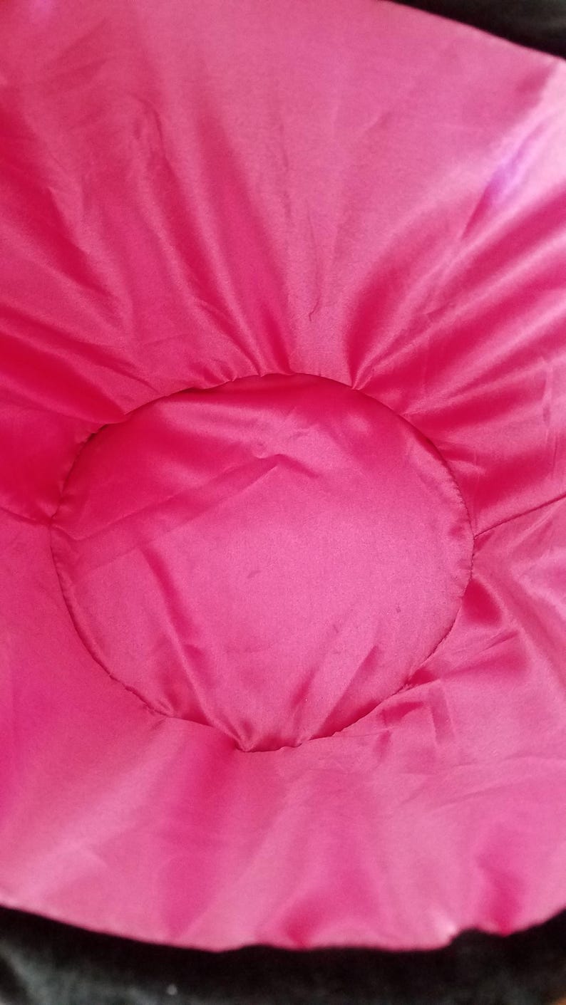 Puede incluir: Primer plano de un elemento de tela rosa brillante, circular, posiblemente un coj&iacute;n o parte de un textil m&aacute;s grande. La tela tiene una textura suave y ligeramente brillante, con un borde m&aacute;s oscuro visible en la parte inferior.