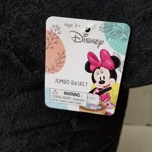 Puede incluir: Una cesta jumbo de felpa negra con una etiqueta de Disney con Minnie Mouse. La etiqueta incluye el texto "Age 3+" y una advertencia de peligro de asfixia. La cesta est&aacute; dise&ntilde;ada para ni&ntilde;os.