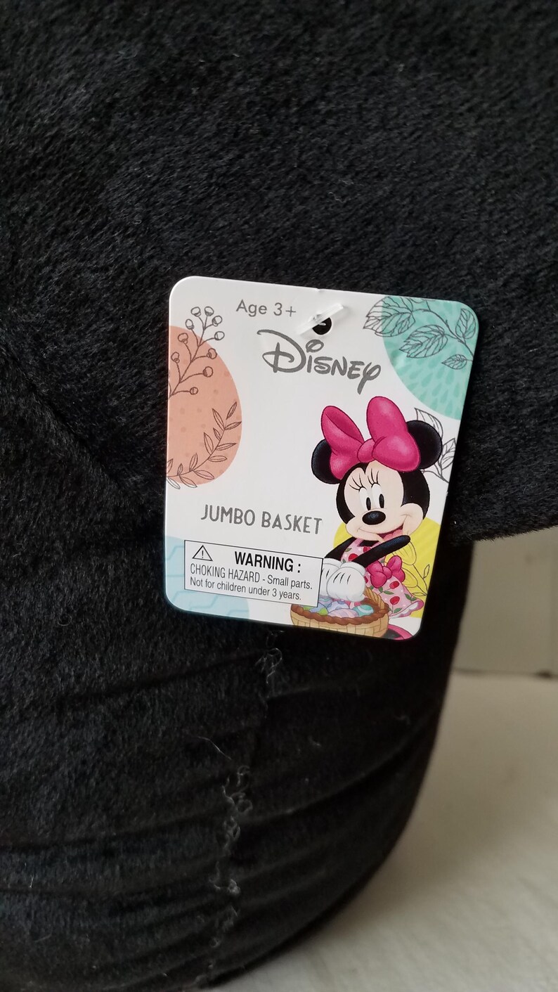 Puede incluir: Una cesta jumbo de felpa negra con una etiqueta de Disney con Minnie Mouse. La etiqueta incluye el texto "Age 3+" y una advertencia de peligro de asfixia. La cesta est&aacute; dise&ntilde;ada para ni&ntilde;os.