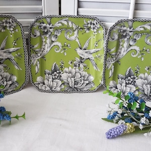 Puede incluir: Tres platos cuadrados decorativos con fondo verde lima y diseño floral y de pájaros en blanco y negro. Los platos tienen un borde de cadena en blanco y negro. Flores artificiales en primer plano.