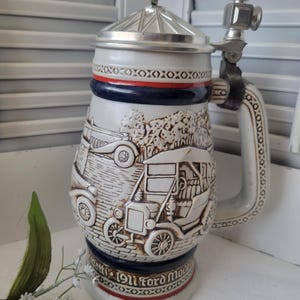 May include: A vintage ceramic beer stein with a silver lid and handle. The stein features embossed images of antique cars and the text "Battle 1911 Ford Model". The design includes red and blue accents.