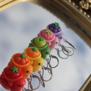 May include: A set of six colorful turtle earrings, each with a different fruit design on their shells. The fruits include a tomato, orange, green apple, strawberry, and grapes. The earrings are made of a glossy material and have silver-colored ear wires.