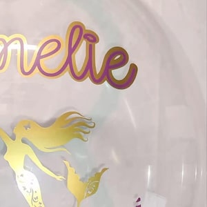 Può includere: Un palloncino a bolla trasparente riempito con palloncini metallici blu, oro e viola. Il palloncino presenta una silhouette di sirena dorata, una stella marina dorata e il nome "Amelie" in oro. È visibile anche il numero 6 viola.