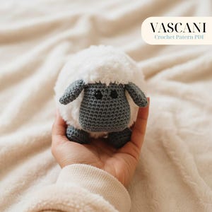 May include: A handmade crochet sheep toy with a fluffy white body and grey face, ears, and feet. The sheep has black safety eyes. The image includes the text "VASCANI Crochet Pattern PDF".